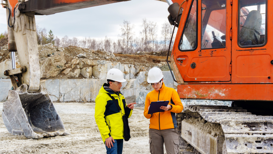 Two construction workers with white hard hats are looking at a smart tablet together in front of a construction site